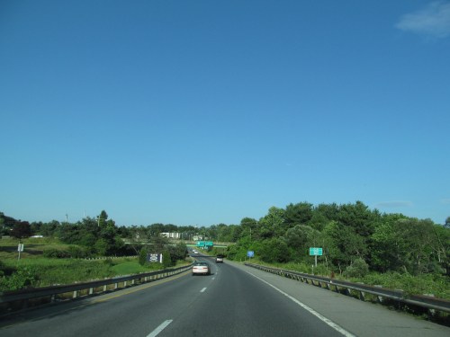 A clear highway stretches ahead, flanked by greenery under a bright blue sky. A car drives towards the horizon.