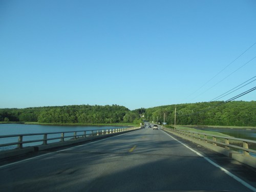 A scenic view of a road and bridge alongside a river, surrounded by lush green trees under a clear blue sky.