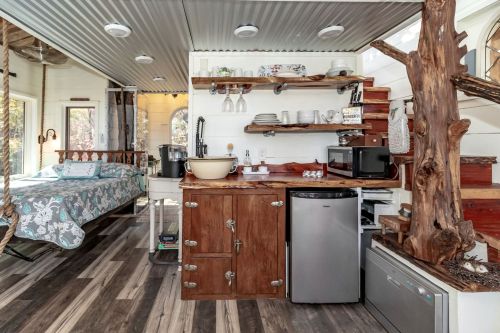 Cozy interior of a tiny home featuring a kitchen area, dining space, and a bed with floral bedding. Natural wood accents.