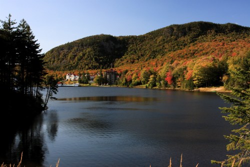 A serene lake surrounded by colorful autumn foliage and mountains, with a building visible in the background.