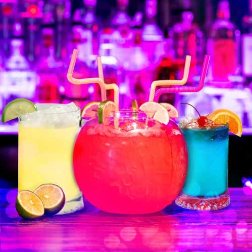 Three colorful cocktails with garnishes, set against a vibrant bar backdrop with purple lighting.