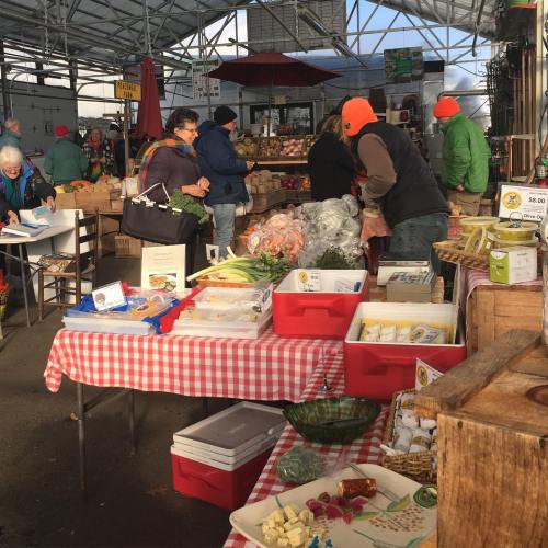A bustling farmers market with vendors and shoppers, featuring fresh produce and colorful displays on checkered tables.