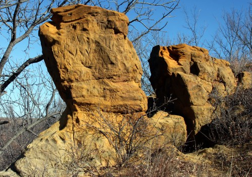 Two large sandstone rock formations with visible textures, surrounded by sparse vegetation and a clear blue sky.