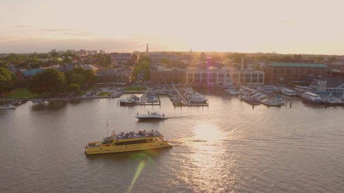 A yellow ferry navigates a calm river at sunset, with a marina and buildings in the background.