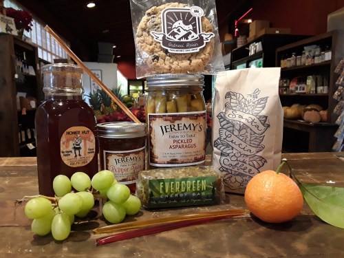 A collection of local products: honey, pickled asparagus, granola, coffee, grapes, a tangerine, and an energy bar on a wooden table.