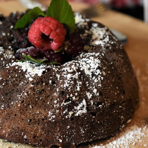 A chocolate bundt cake topped with raspberries and mint, dusted with powdered sugar on a wooden surface.