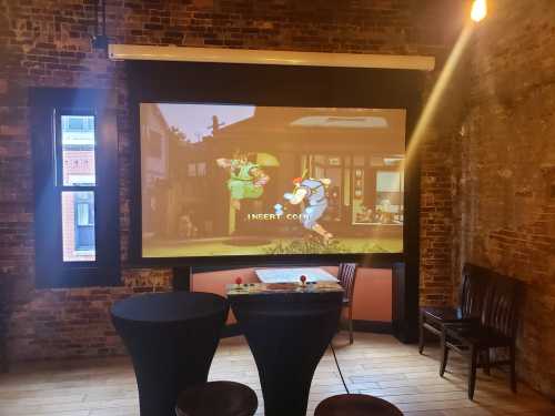 A large screen displays a colorful arcade game in a rustic room with two black tables and wooden chairs.