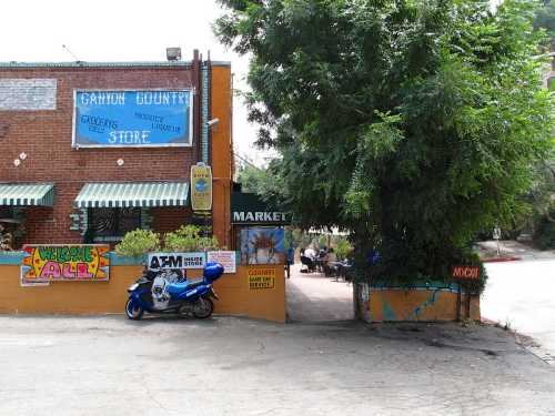 A colorful storefront of a grocery store with a blue motorcycle parked outside and trees nearby.