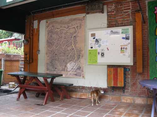 A dog stands near a wall with a large map and a bulletin board, surrounded by outdoor seating.