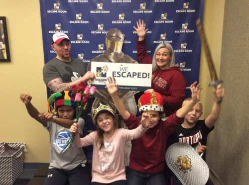 A group of six people celebrating with props and a sign that says "We Escaped!" in front of an escape room backdrop.