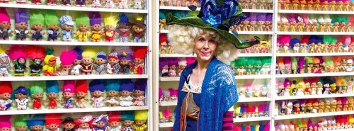 A person in a colorful outfit stands beside a wall of vibrant troll dolls in various styles and colors.