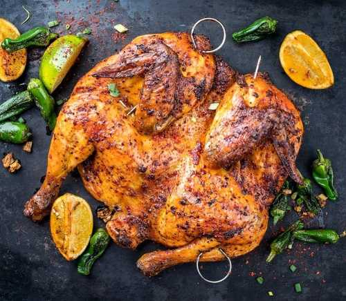 A beautifully roasted chicken with crispy skin, garnished with lime, lemon, and green peppers on a dark surface.