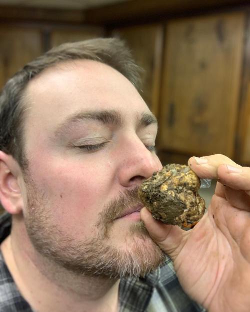 A man with a beard closes his eyes while holding a large, round, textured food item close to his nose.