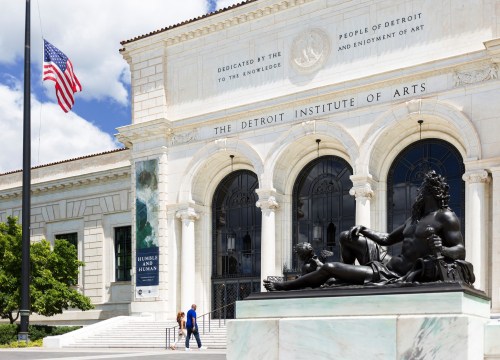 The Detroit Institute of Arts building with a statue in front, featuring a blue sky and people walking by.