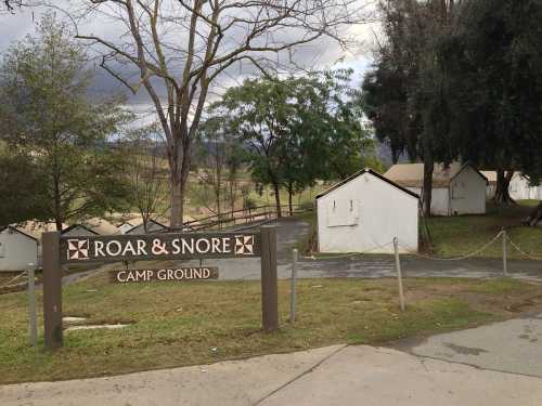 Sign for "Roar & Snore Camp Ground" with white tents and trees in the background under a cloudy sky.