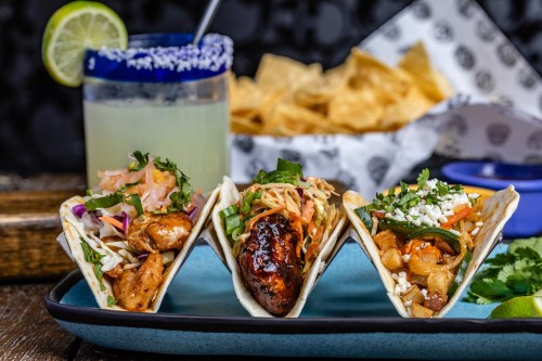 Three colorful tacos filled with various ingredients, served with a margarita and tortilla chips in the background.