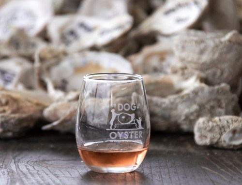 A glass of rosé wine sits in front of a pile of oyster shells, featuring a logo that reads "Dog Oyster."