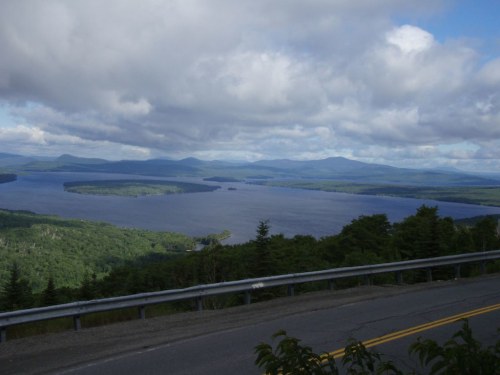 A scenic view of a lake surrounded by green forests and mountains under a cloudy sky.