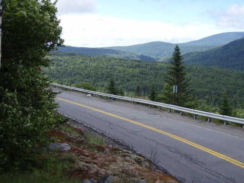 A winding road curves through a lush green landscape with rolling hills and a cloudy sky in the background.