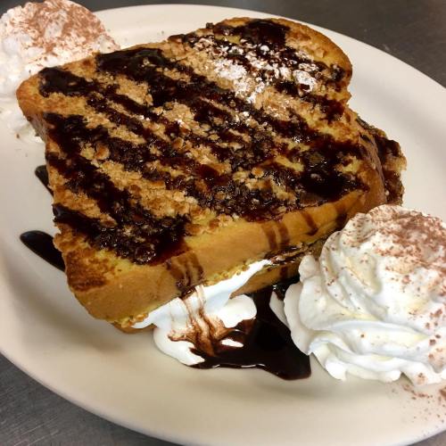 A slice of French toast drizzled with chocolate sauce, topped with whipped cream and sprinkled with powdered sugar.