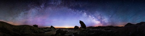 A panoramic view of a starry night sky with the Milky Way over rocky terrain and a colorful horizon.