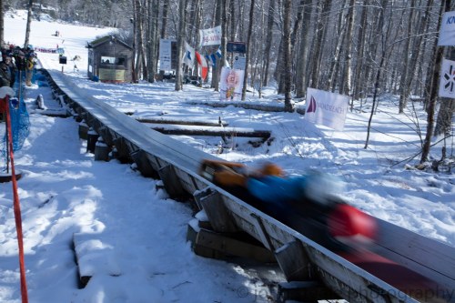 A person sledding down a snowy track, surrounded by trees and spectators in winter attire.