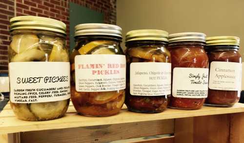 Five jars of pickles and preserves on a wooden shelf, labeled with flavors like sweet pickles and jalapeño hot pickles.