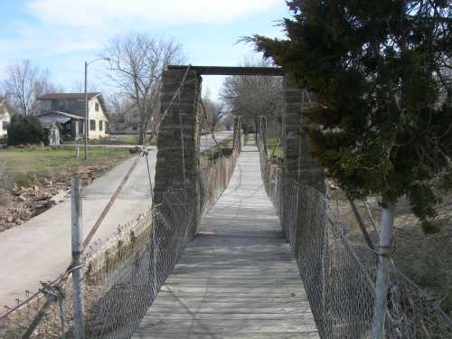 A wooden suspension bridge stretches over a small creek, surrounded by trees and a house in the background.