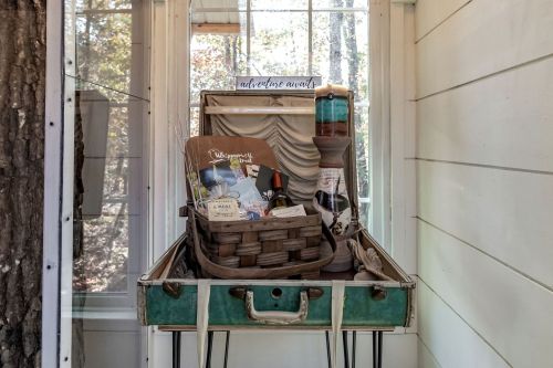 A vintage suitcase filled with a basket, decor items, and a sign, set against a backdrop of trees and a glass wall.