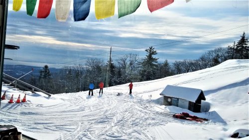 Three skiers on a snowy slope, with colorful prayer flags in the foreground and trees in the background.