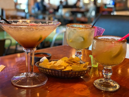 Three colorful cocktails on a table with a basket of tortilla chips in a lively restaurant setting.
