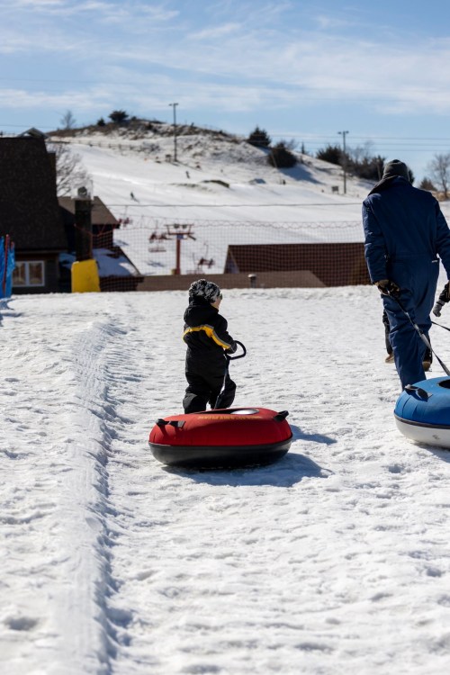 A child in a yellow and black snowsuit pulls a red sled while walking on snow, accompanied by an adult in blue.