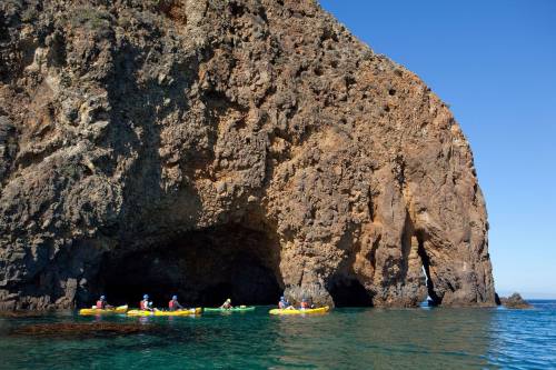 A group of kayakers paddles near a large rocky cliff with a cave opening, surrounded by clear blue water.