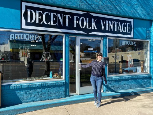 A woman stands outside a bright blue vintage shop, "Decent Folk Vintage," with a welcoming gesture.