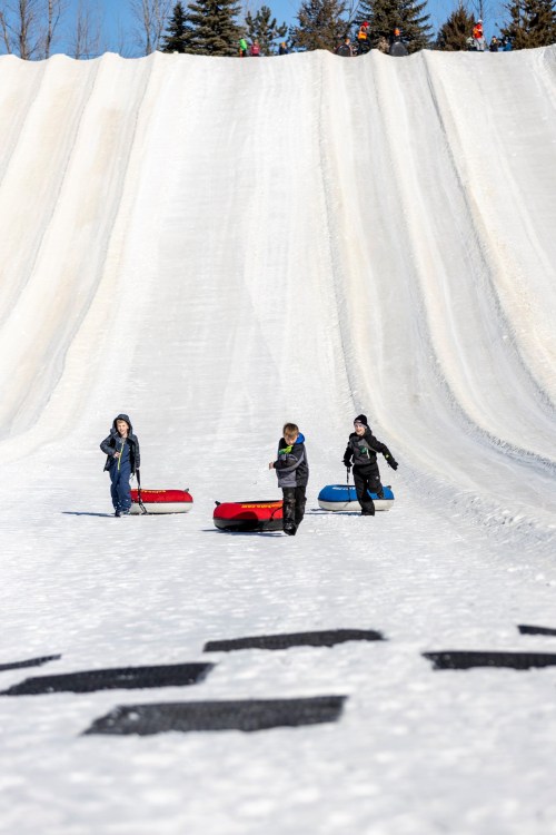 Three children walk up a snowy hill, pulling colorful sleds behind them on a sunny winter day.