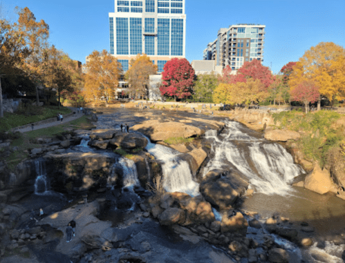 A scenic view of waterfalls surrounded by autumn trees and a city skyline in the background. People enjoy the area.