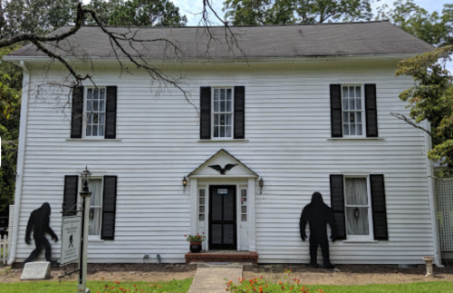A white two-story house with black shutters, featuring two large silhouettes of Bigfoot on either side of the entrance.