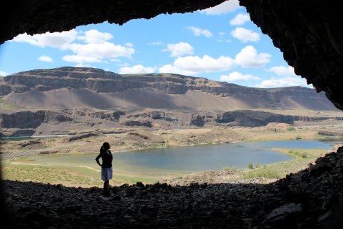 A person stands in a cave, overlooking a scenic landscape with a lake and mountains under a blue sky with clouds.
