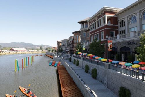 A scenic waterfront with colorful umbrellas, kayaks, and modern buildings along a calm river.