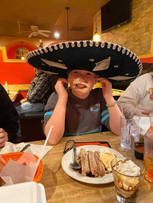 A child wearing a large sombrero, smiling with whipped cream on their face, sitting at a restaurant table with desserts.