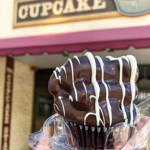 A close-up of a chocolate-covered cupcake with white drizzle, held in a hand outside a bakery.