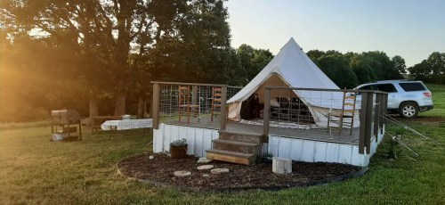 A glamping tent on a wooden deck in a grassy field, with a grill and a car nearby, under a clear sky.