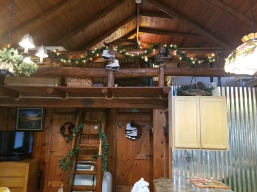 Cozy cabin interior with wooden beams, holiday decorations, and a loft area, featuring a rustic kitchen and warm lighting.
