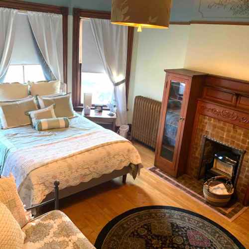 Cozy bedroom with a bed, decorative pillows, a radiator, and a fireplace, featuring large windows and warm lighting.