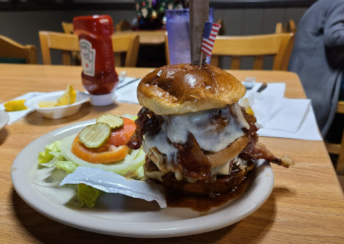 A large burger with bacon, cheese, and a flag on top, served with lettuce, tomato, pickles, and ketchup on the side.