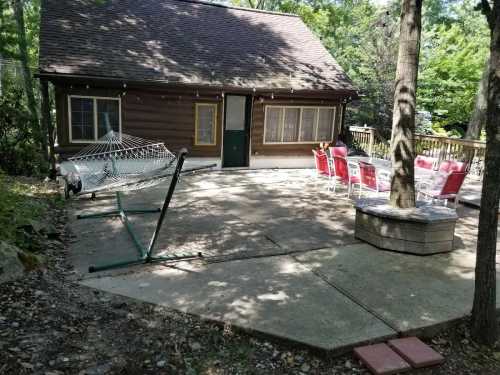 A cozy cabin surrounded by trees, featuring a hammock, patio furniture, and a fire pit on a concrete patio.