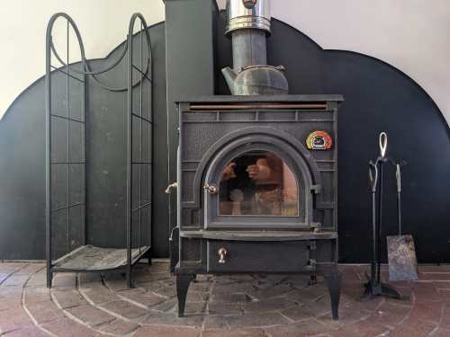 A black wood-burning stove with a metal chimney, next to a firewood rack and fireplace tools on a tiled floor.