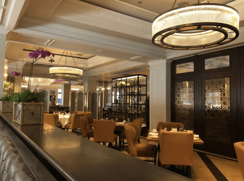 Elegant restaurant interior with wooden tables, yellow chairs, and decorative lighting. A touch of purple orchids adds color.