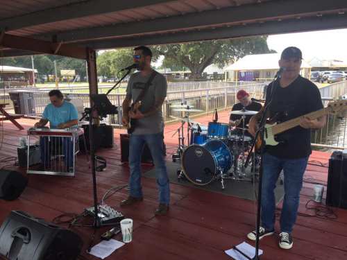 A band performs on a wooden stage by the water, featuring a singer, guitarist, drummer, and steel guitarist.