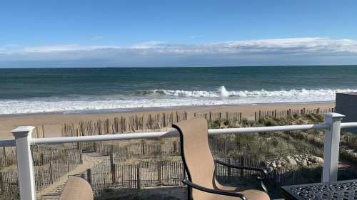 A serene beach view with gentle waves, sandy shore, and a clear blue sky, framed by a railing and a chair.